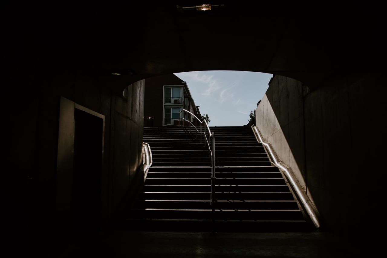 Looking out from a dark concrete underpass towards a broad flight of steps climbing to street level, twin metal handrails catching the light, a building and pale sky visible at the top