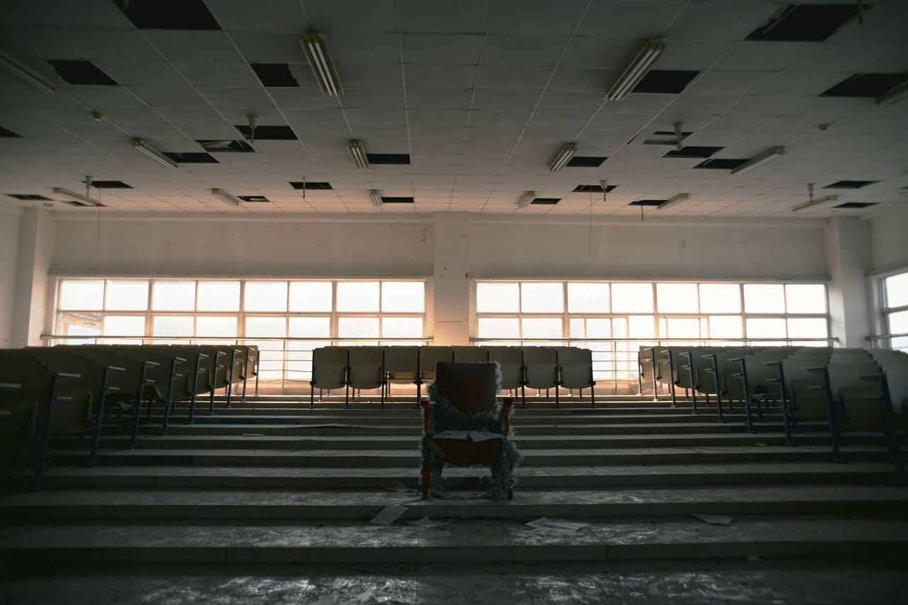 A large empty lecture hall photographed straight down its centre line, rows of vacant seats and fixed desks stepping away in symmetry, lit coldly from high side windows with deep shadow between the ranks.