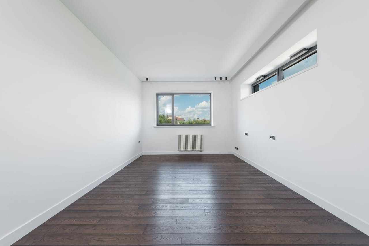 An entirely empty apartment room with plain walls and a bare parquet floor, daylight entering from a tall window on one side; no furniture and nothing to give the space a sense of scale
