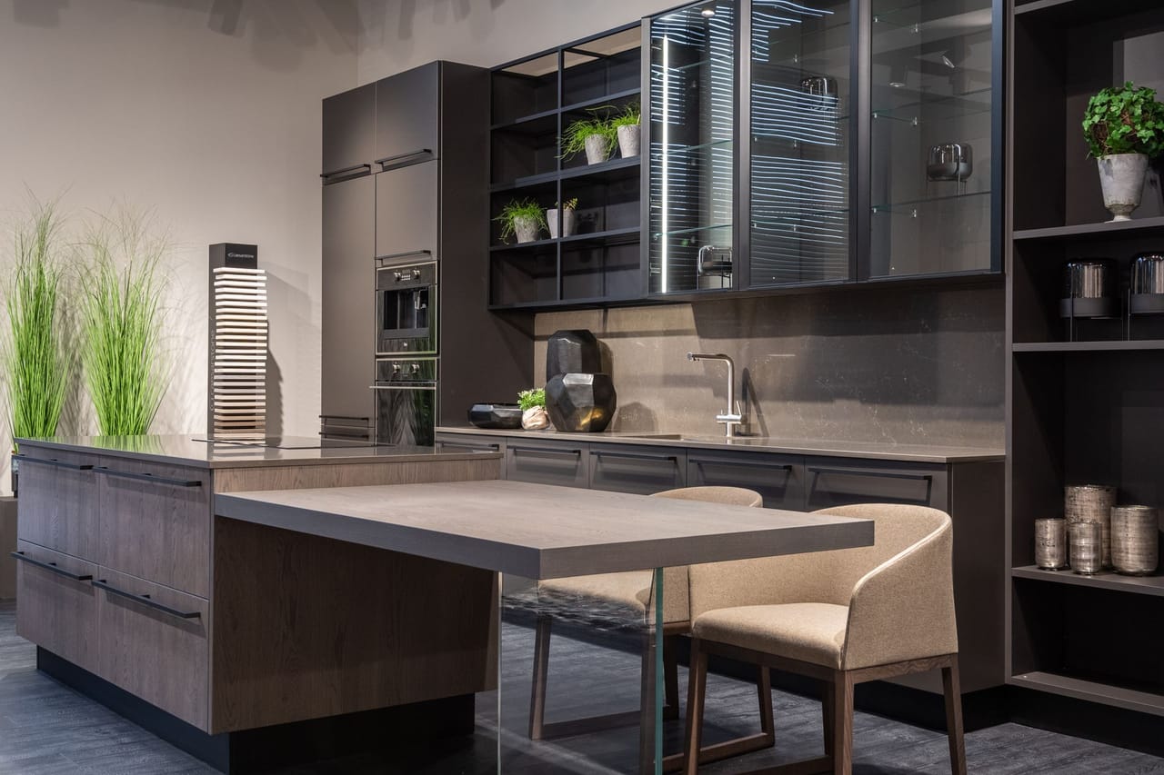 A kitchen in matte charcoal cabinetry with glass-fronted upper units lit from inside, a stone worktop and splashback, and a wooden island table with two pale upholstered chairs.