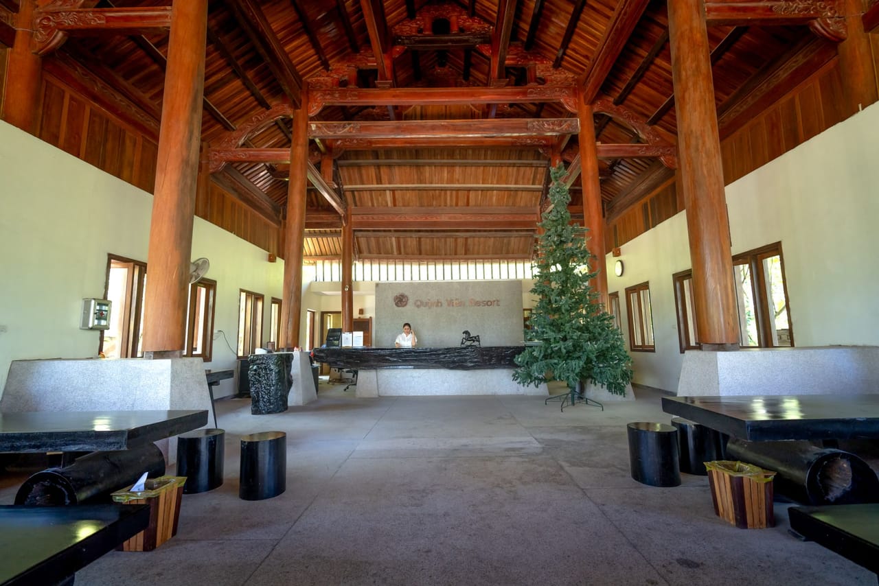 A long unstaffed reception counter set against a panelled wall, standing under a high vaulted ceiling crossed by dark timber beams, with no one behind or in front of it
