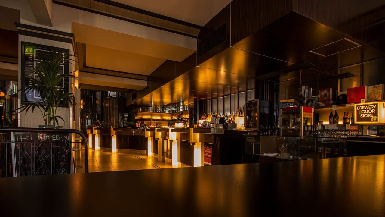 The interior of a bar after dark with no one in it: an unoccupied counter running down the frame, stools pushed in, and the bottle shelving behind lit from within so the whole back wall glows against the dark ceiling.