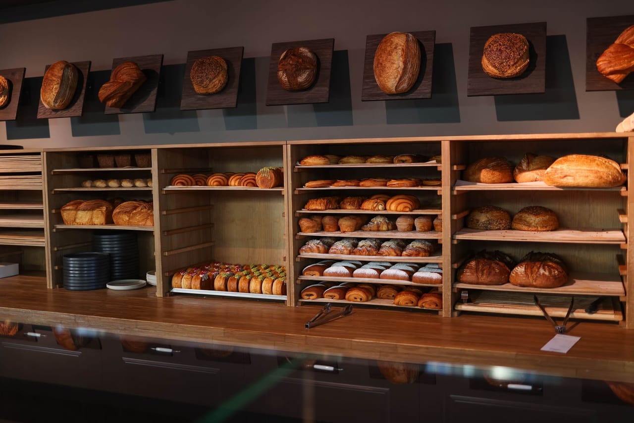 Behind a long wooden counter, open timber shelving is stacked with round sourdough loaves, pastries and rolls on wire trays, with single loaves mounted on dark wooden boards along the wall above