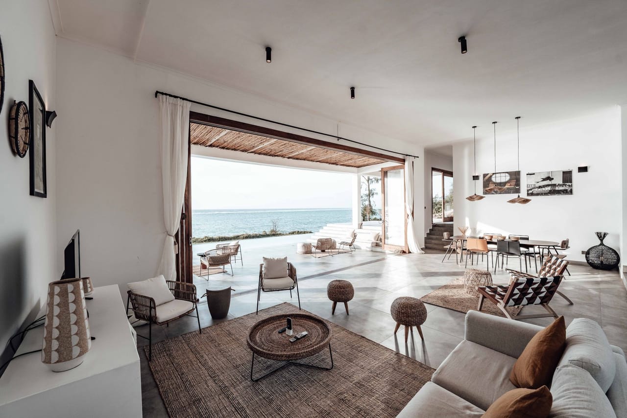 A sparsely furnished living room with pale walls, its wide opening framing flat open sea and horizon beyond